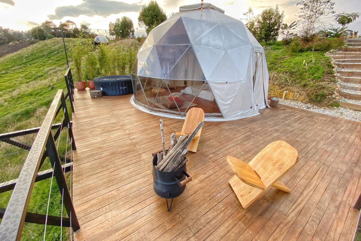 Une tente en forme de dôme géodésique dotée d’un toit transparent, d’une terrasse en bois, d’un foyer et d’un espace détente avec une chaise et un banc en bois.