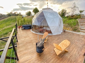 A geodesic dome tent with a transparent roof, a wooden deck, a fire pit, and a seating area with a wooden chair and a bench.
