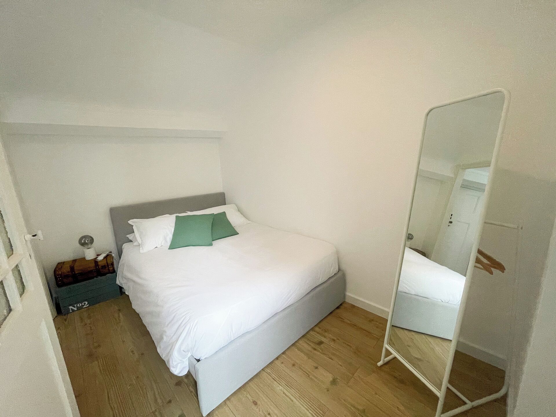 Una cama bien hecha con ropa blanca y dos almohadas verdes, piso de madera y un armario blanco con espejo.