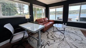 A modern living room with a brown leather sofa, a white desk, and a small round table. Large windows offer views of a lake and forest.