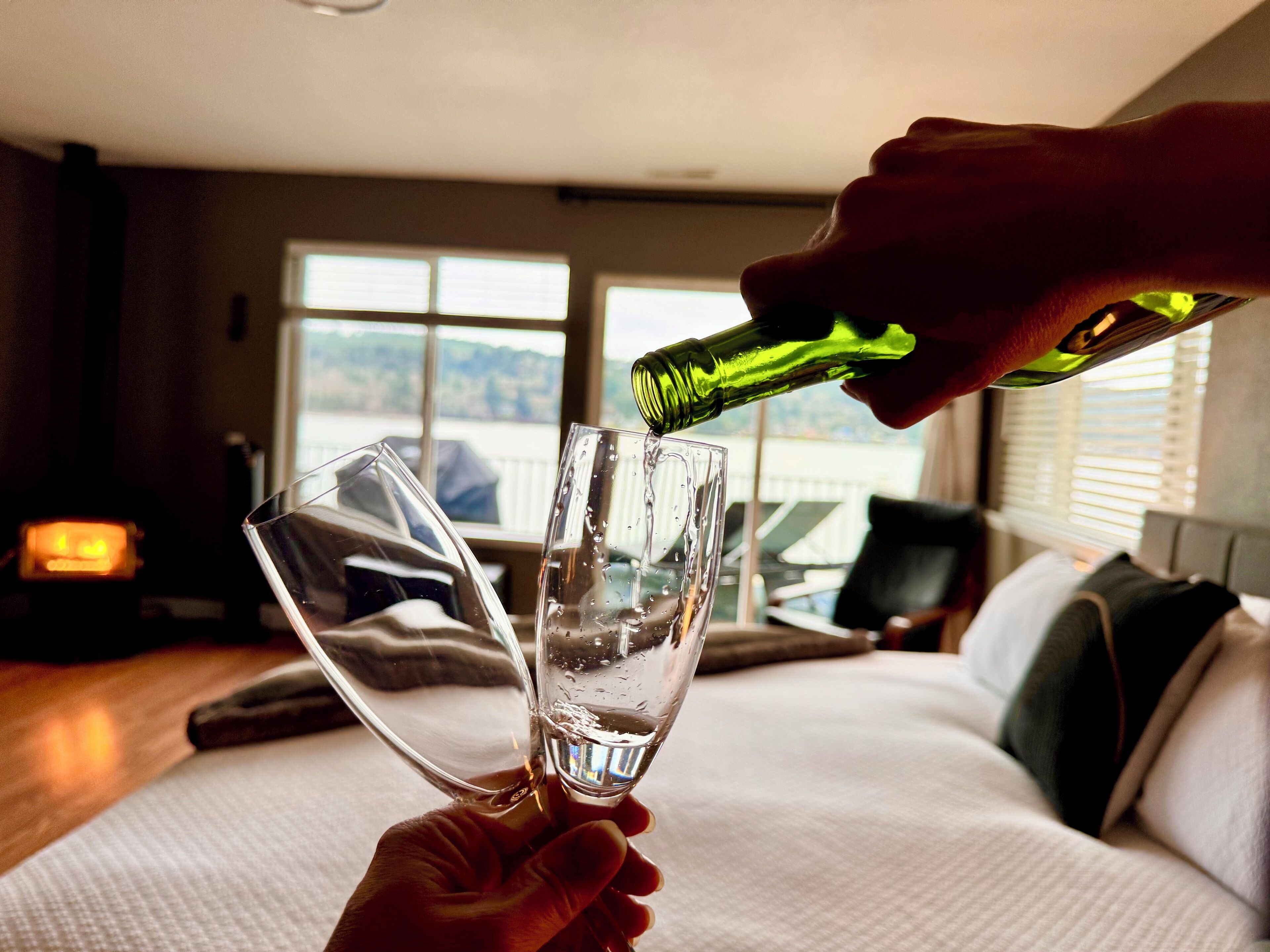 Een hand schenkt water in een glas op een bed in een kamer met een groot raam en een open haard.