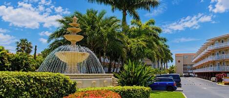 A fountain with multiple tiers, surrounded by well-maintained hedges and flowering plants, with a clear blue sky and a building in the background.