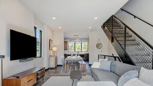 A modern living room with a flat-screen TV, a sofa, a dining area, and a staircase. - Downtown WP Loft 21 I Rooftop Hot Tub & Lounge I Great Views I Bunks I Discounted Attractions (Winter Park)