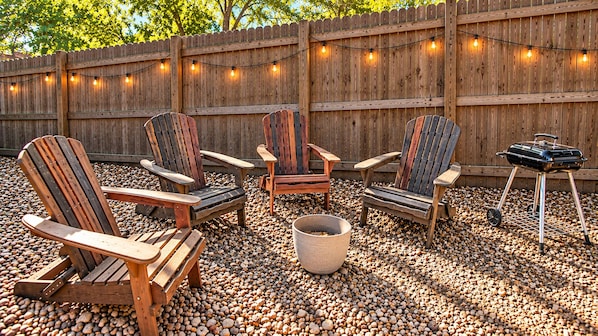 An outdoor seating area with Adirondack chairs, a barbecue grill, and string lights.