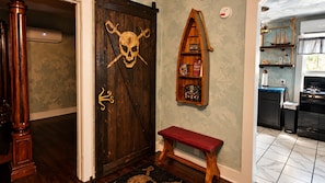 Una puerta de madera con calavera y tibias cruzadas, un estante con libros, un banco y una zona de cocina con estufa.