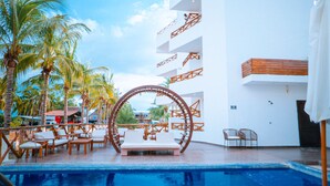A pool area with lounge chairs and a wooden archway, surrounded by palm trees and a white building with balconies. - Baos Hotel & Restaurant (Aticama)