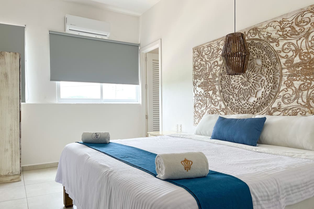 A hotel room with a large bed, a decorative headboard, an air conditioning unit, and a window with blinds.
