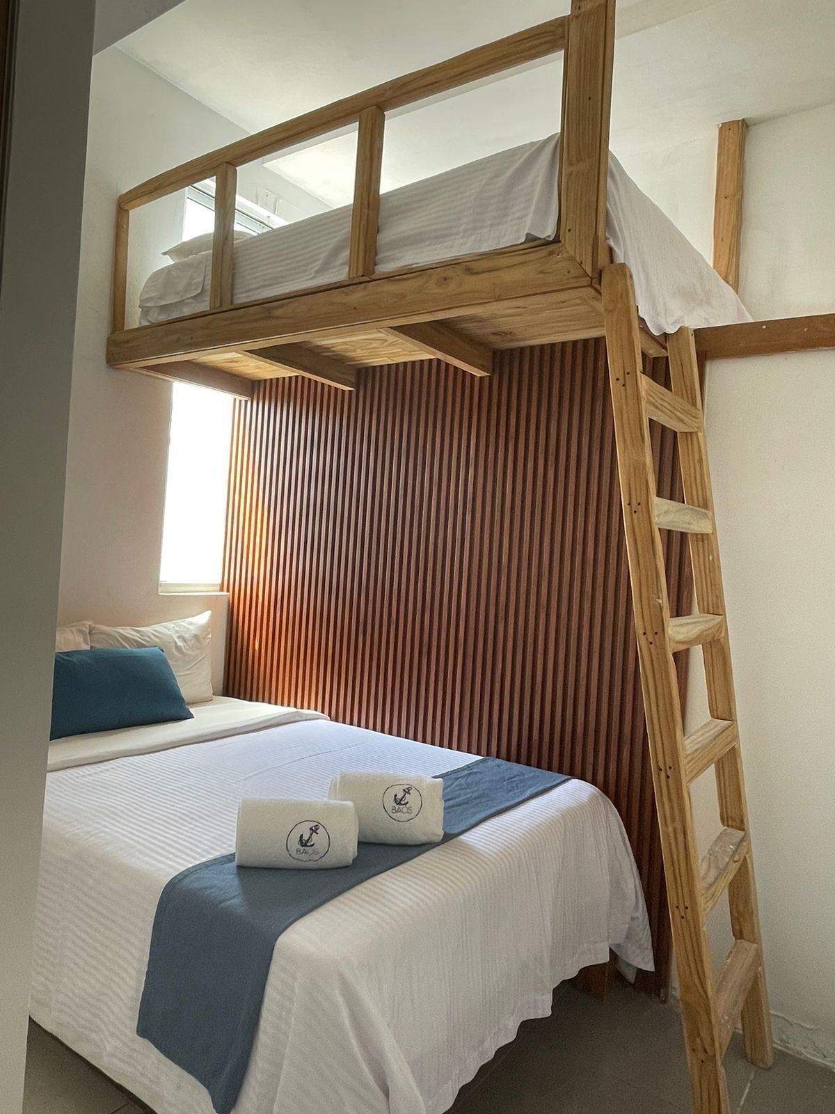 A bunk bed with a wooden ladder, a window with blinds, and a bed with a blue pillow and a white blanket.