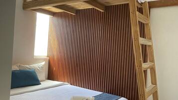 Una cama individual con escalera de madera, una ventana con persianas, y una cama con almohada azul y manta blanca.