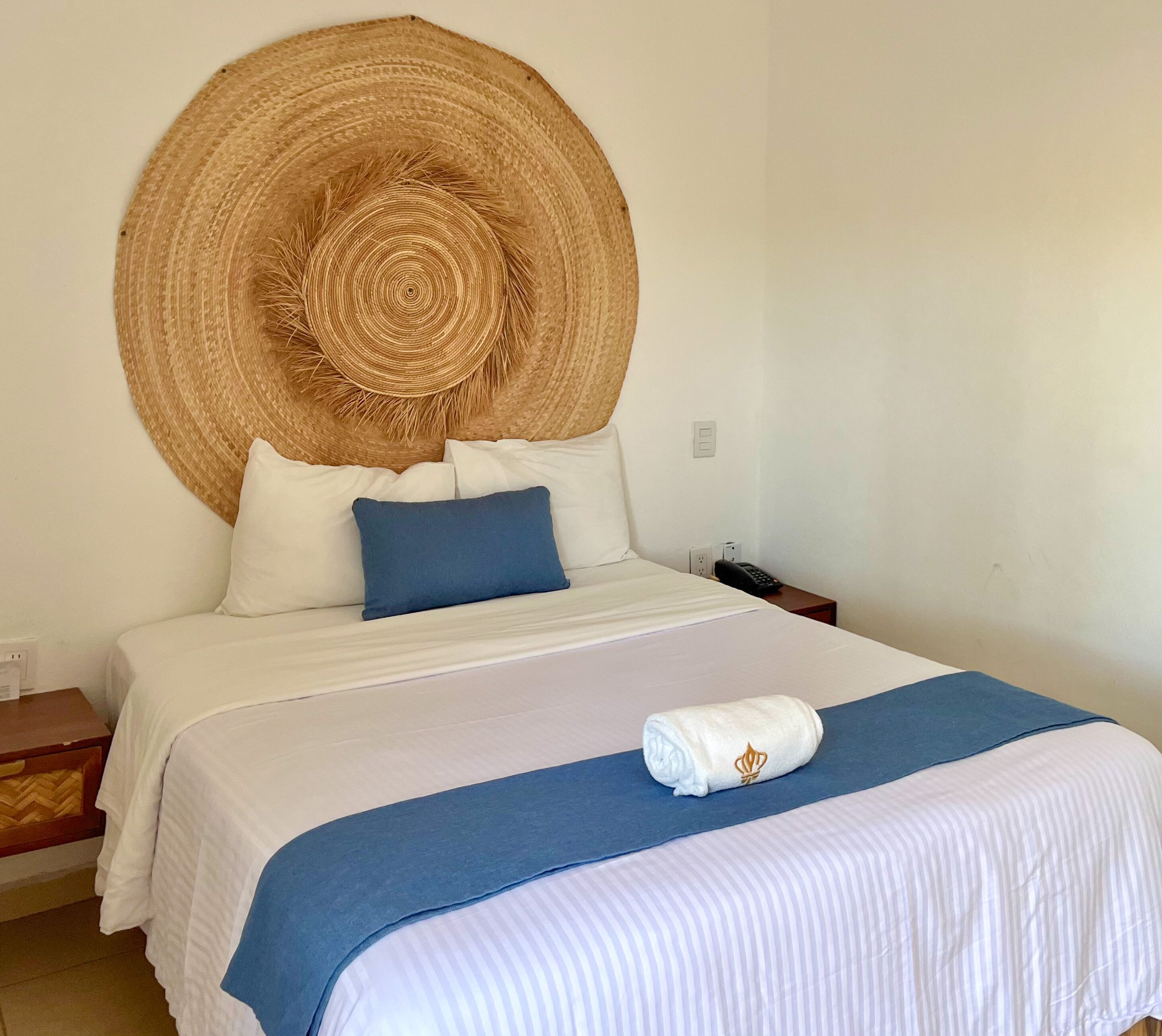 Cama bem arrumada com roupa de cama branca e azul, cabeceira grande trançada e toalha branca com um logotipo.