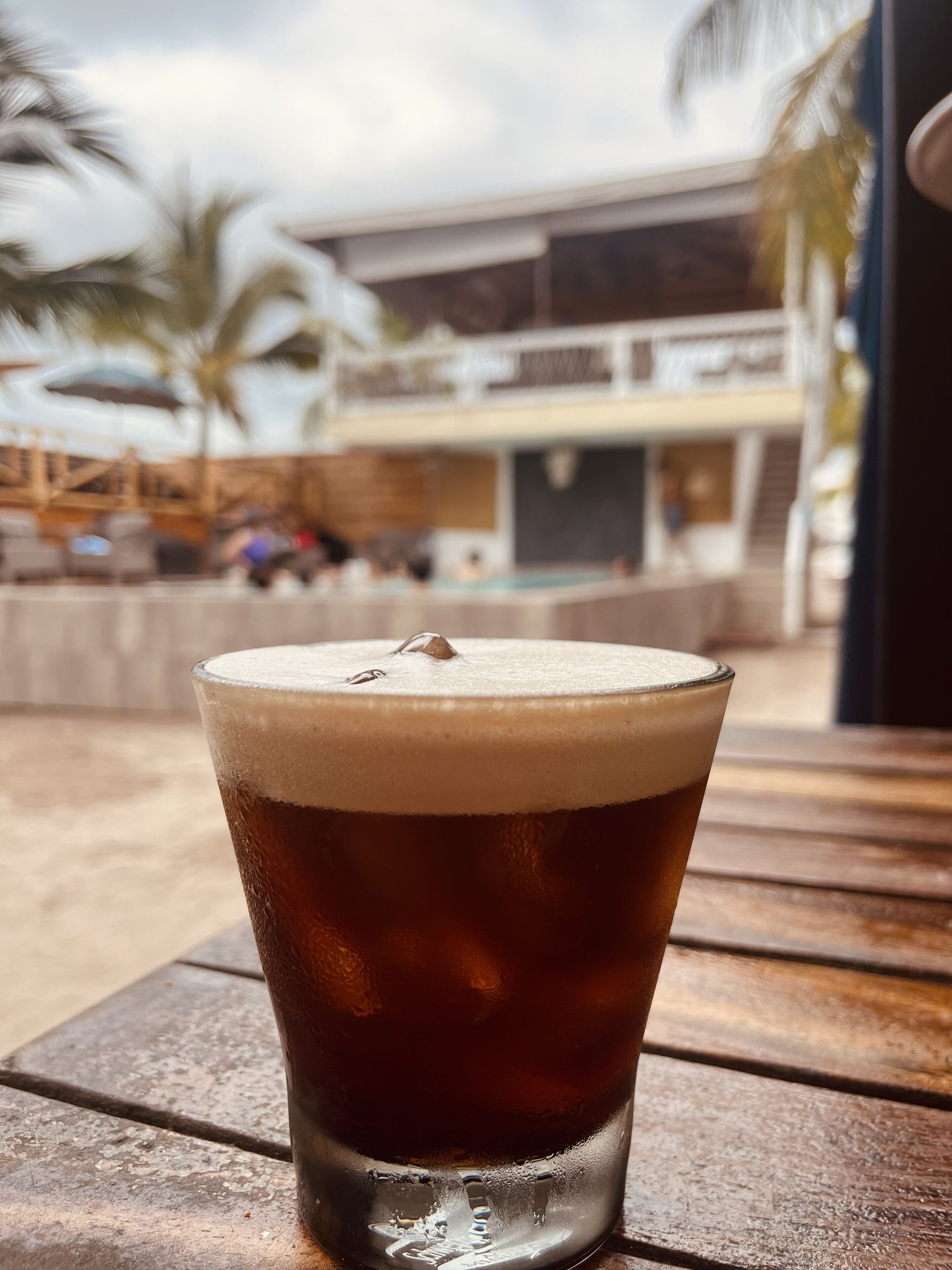 A glass of beer on a wooden table with a view of a building and palm trees in the background.