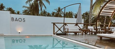 Una piscina con sillas de descanso y sombrillas, rodeada de palmeras.