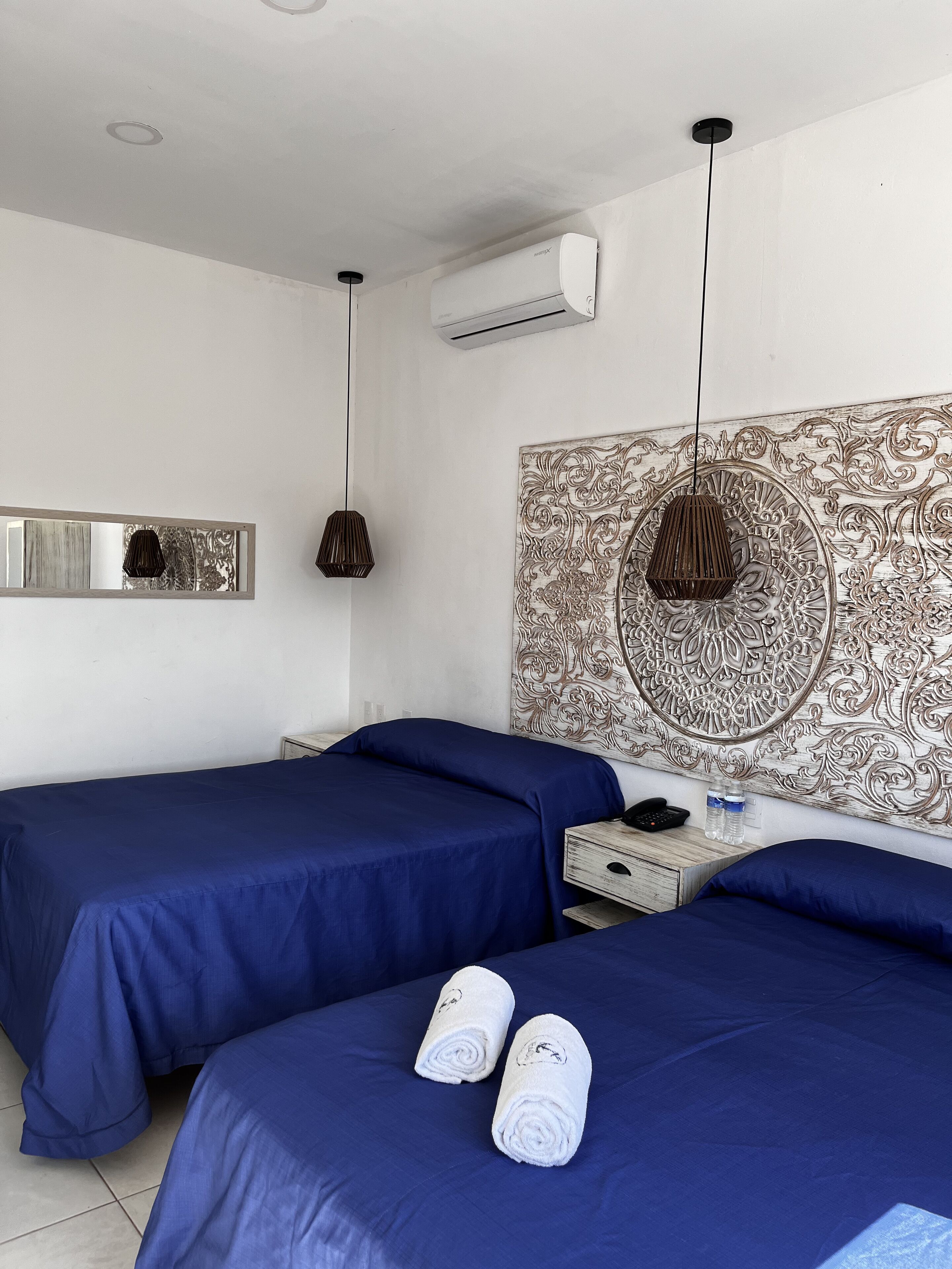 A hotel room with two beds, a large decorative tapestry on the wall, a modern pendant light, and a small bedside table with a phone and water bottles.