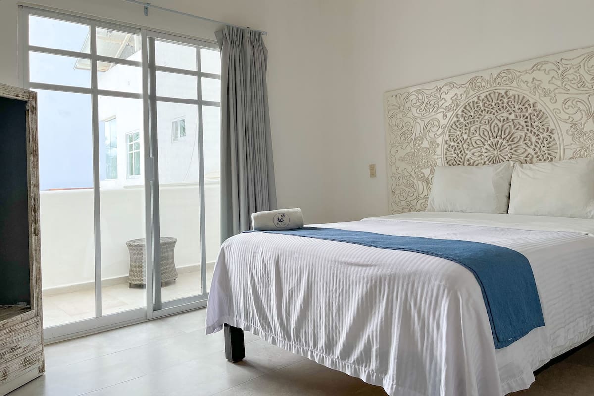A hotel room with a large bed, a decorative headboard, a view through a sliding glass door, and a small table with a chair.