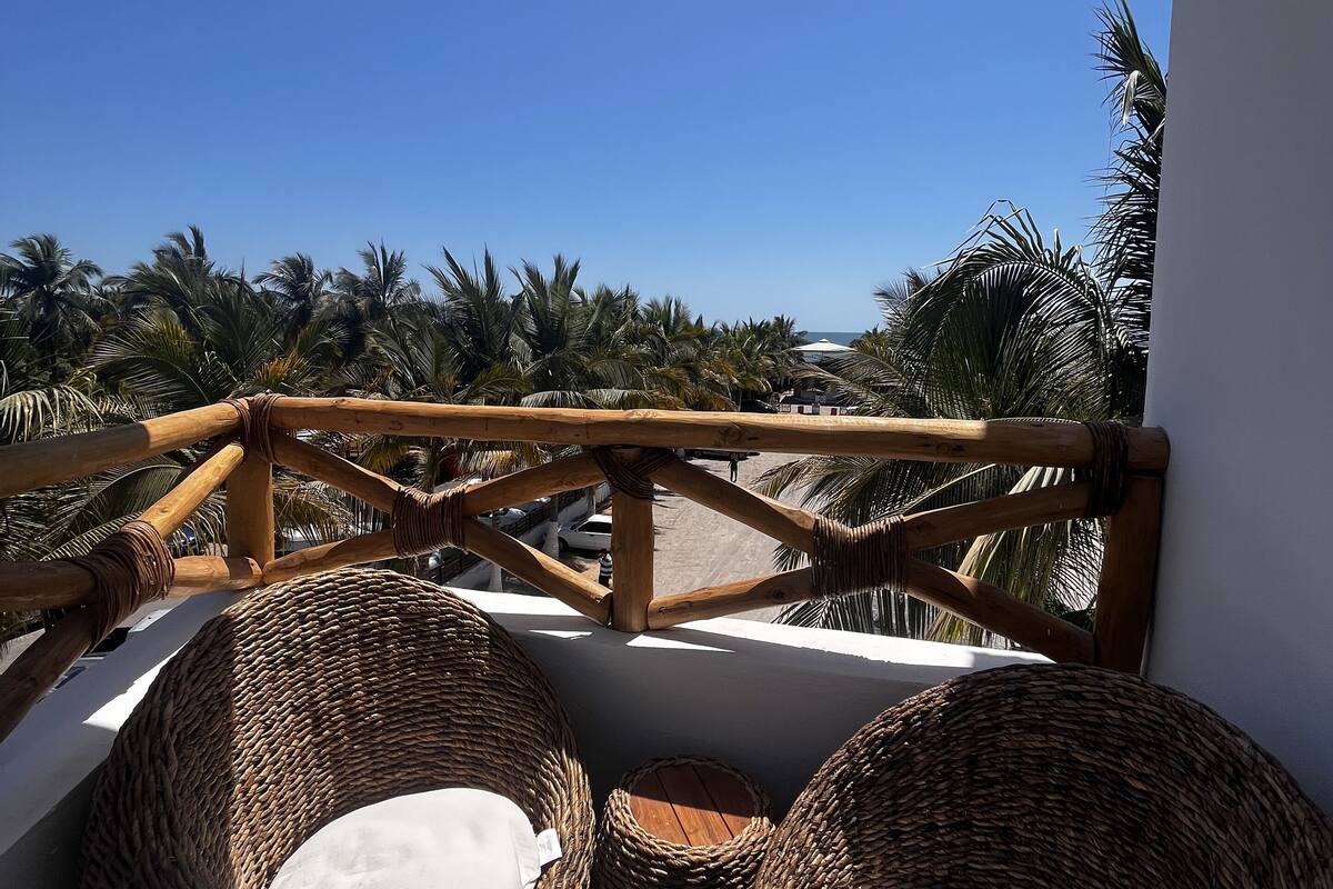 A balcony with wicker chairs and a wooden railing overlooking palm trees.