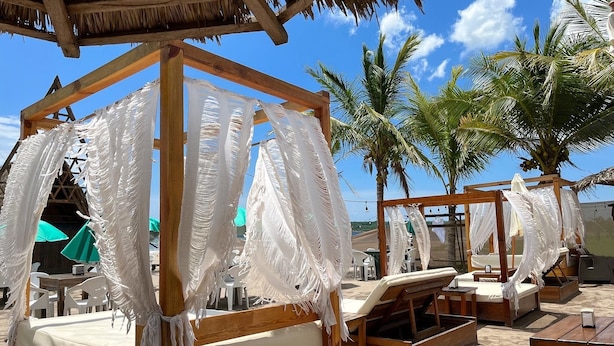 A beach cabana with a wooden frame and white fabric canopy, surrounded by palm trees and beach chairs. - Baos Hotel & Restaurant (Aticama)