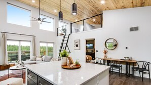 A spacious living area with a large island, a loft bedroom with a balcony, and a dining area with modern furniture. - Mountain & Pond Views, Hot Tub, Stargazing, Kid + Dog Friendly, Loft+3BR (Florissant)
