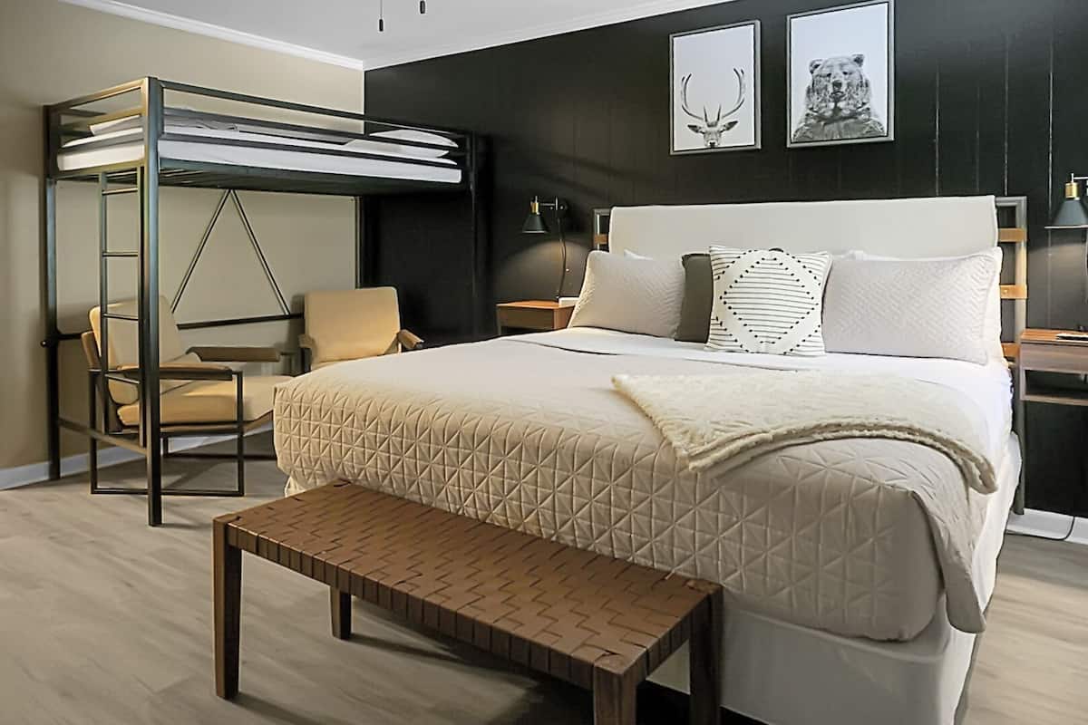 Ein modernes Hotelzimmer mit einem Etagenbett, einem Einzelbett, einem Sessel, einem Nachttisch und einem Beistelltisch. An der Wand hängen zwei gerahmte Bilder.