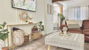 A living room with a television, a brown leather sofa, a patterned rug, and various decorative items on shelves. - Walking distance to Betsie River & 10 minutes to Lake Michigan & Crystal Mnt! (Benzonia)
