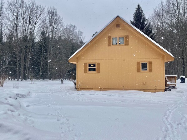 En stuga byggd av trä med ett sadeltak och två fönster, omgiven av snötäckt mark och träd.