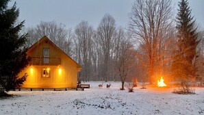 A wooden cabin with a porch, surrounded by a snowy landscape and a fire burning in the distance. - Walking distance to Betsie River & 10 minutes to Lake Michigan & Crystal Mnt! (Benzonia)