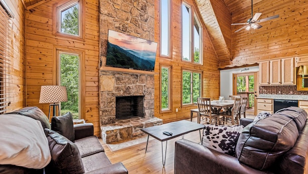 A cozy cabin interior with a stone fireplace, wooden walls, a dining area, and a kitchen. - Mountain Vacation Time! 2 miles to GSMNP@ Line Springs~Hot Tub~Games~2 Decks (Sevierville)