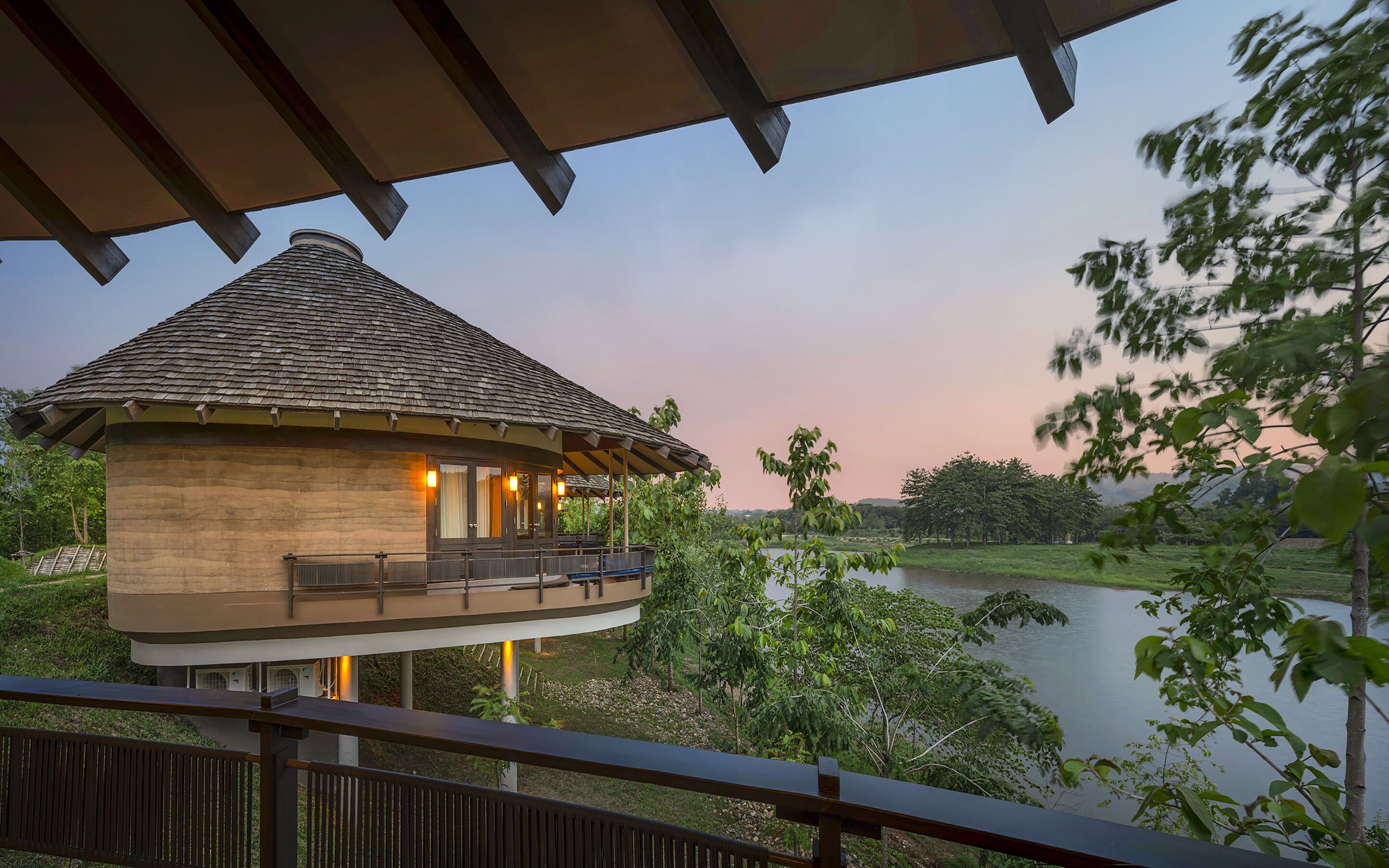 A two-story building with a thatched roof, a balcony with seating, and a view of a lake surrounded by trees.