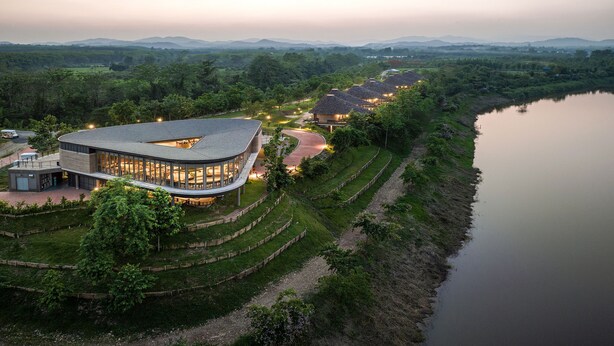 Un hotel moderno con techo curvo y ventanas amplias, ubicado junto a un río con jardines escalonados y vistas a montañas distantes.
