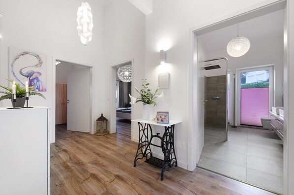 A modern interior with a wooden floor, a black metal table with a vase, a wall-mounted light, and a door leading to a bathroom.