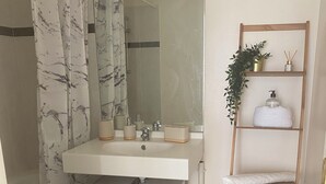 Une salle de bain avec un meuble-lavabo blanc, un miroir, un rideau de douche, une étagère avec des articles de toilette et une petite plante.