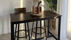 Un coin repas avec une table en bois foncé, deux tabourets en métal noir, un tableau encadré au mur et une plante en pot.