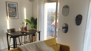 Un salon avec un canapé de couleur moutarde, une table à manger avec des chaises, un luminaire suspendu et une vue dégagée sur l’extérieur grâce à une grande fenêtre.