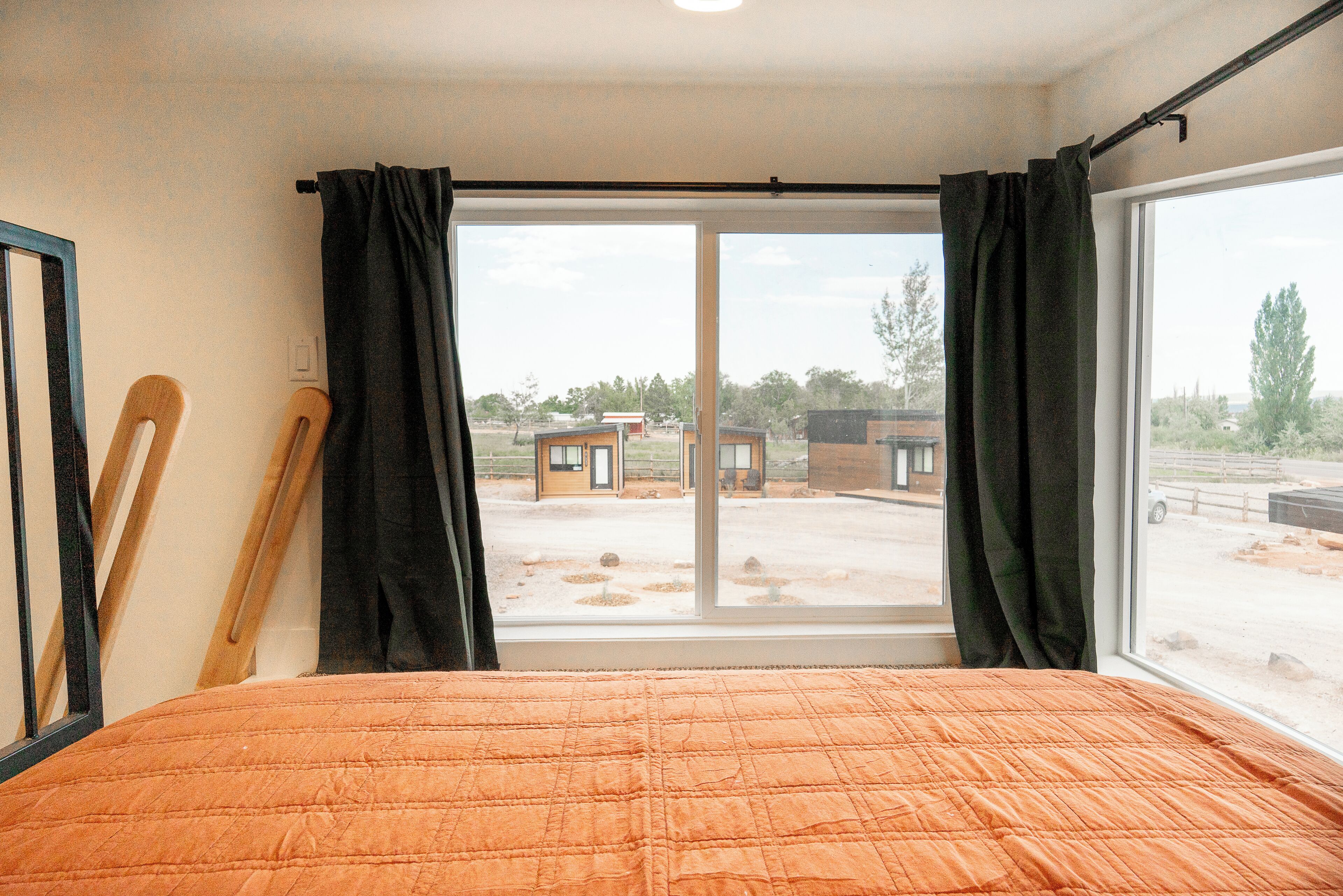 A bedroom with a large window, a bed with an orange quilt, and a view of a building complex outside.
