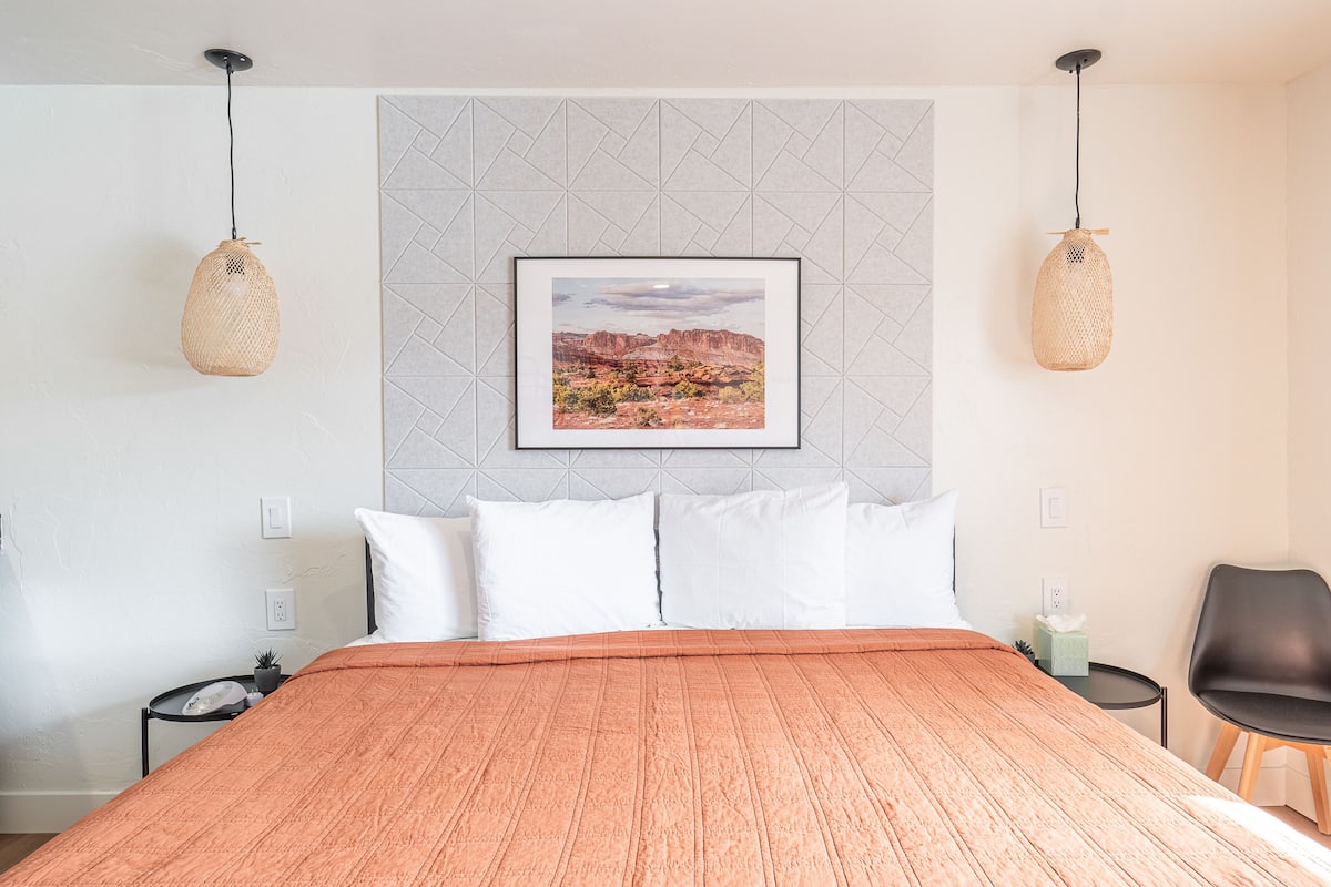 A neatly made bed with a textured orange blanket, a framed landscape picture on the wall, and two hanging wicker lamps.