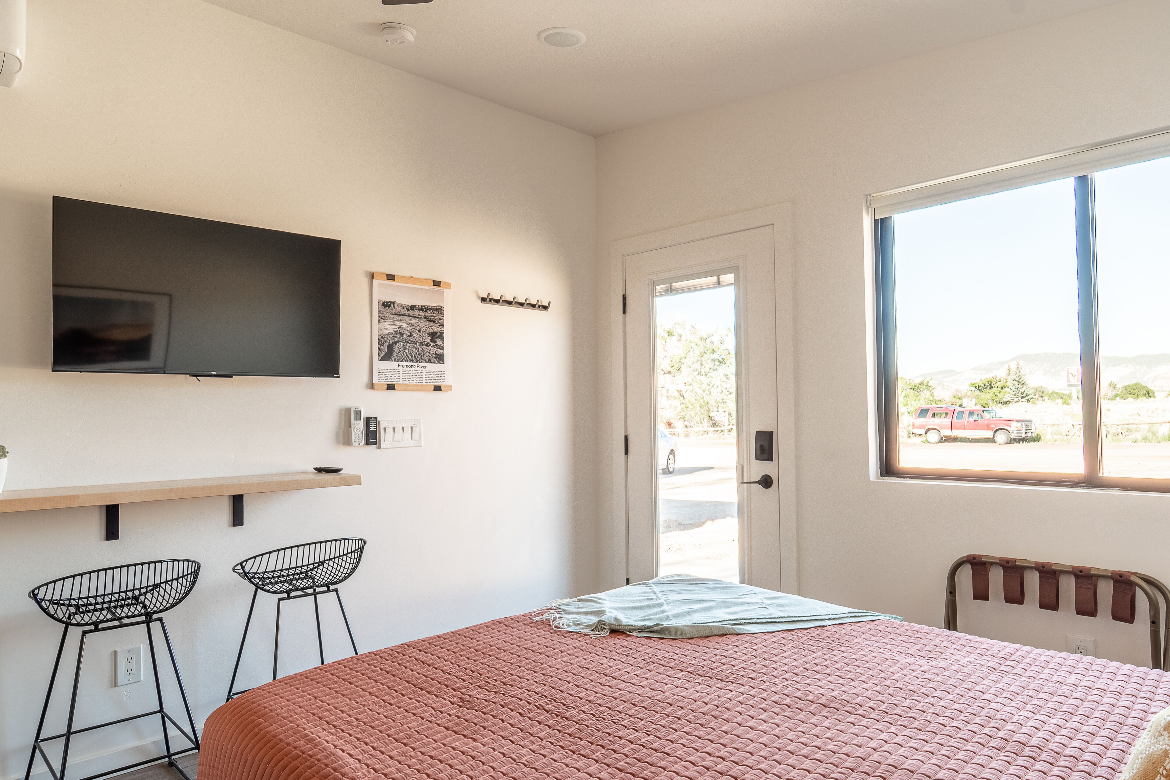 A bedroom with a bed, a TV, two bar stools, a window, and a door.