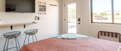 A bedroom with a bed, a TV, two bar stools, a window, and a door.