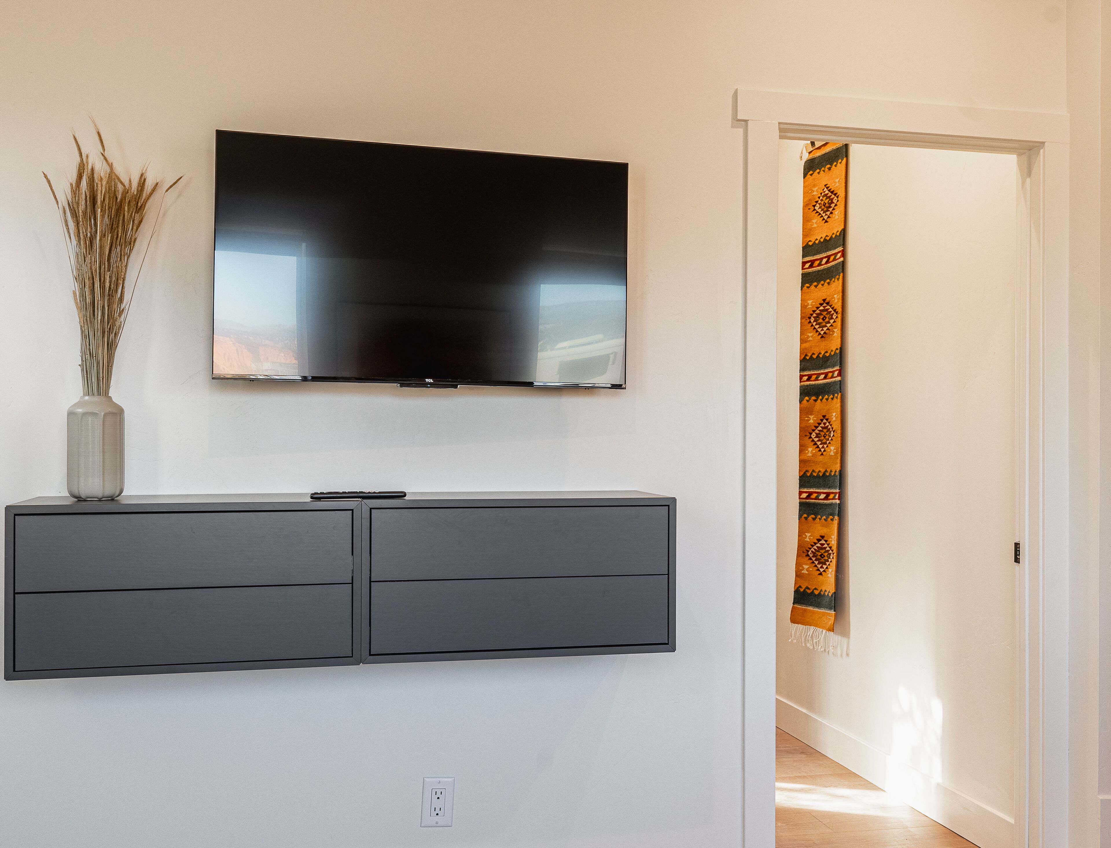 A modern living room with a flat-screen TV mounted on the wall, a grey cabinet below, and a decorative vase with dried plants.