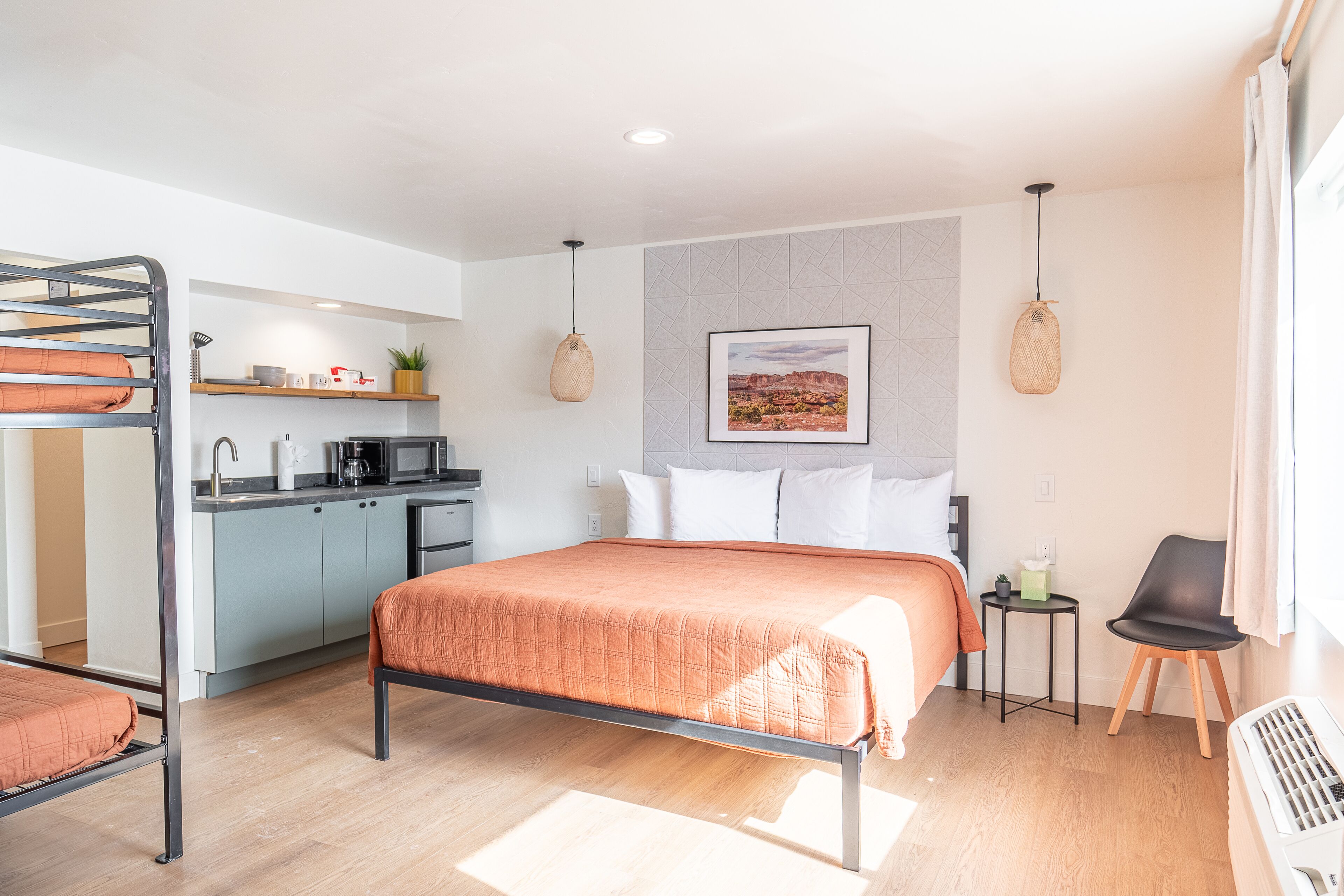 A modern hotel room with a bunk bed, a kitchenette, a bed with an orange blanket, a small table with a chair, and a picture on the wall.