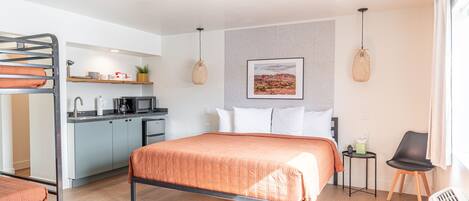 A modern hotel room with a bunk bed, a kitchenette, a bed with an orange blanket, a small table with a chair, and a picture on the wall.