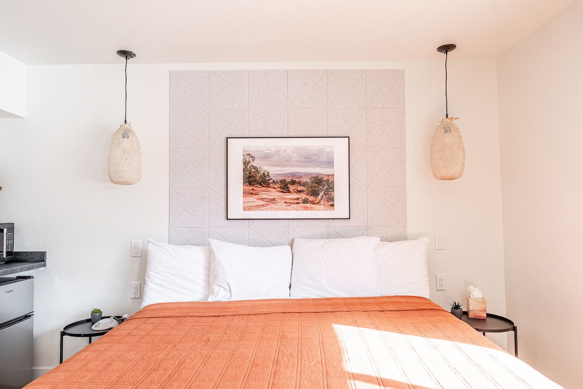 A bedroom with a bed, white pillows, an orange bedspread, a framed picture on the wall, and two hanging lights.