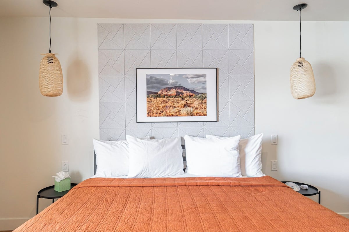 A hotel room with a bed, white pillows, an orange bedspread, a framed picture on the wall, and two hanging wicker lamps.