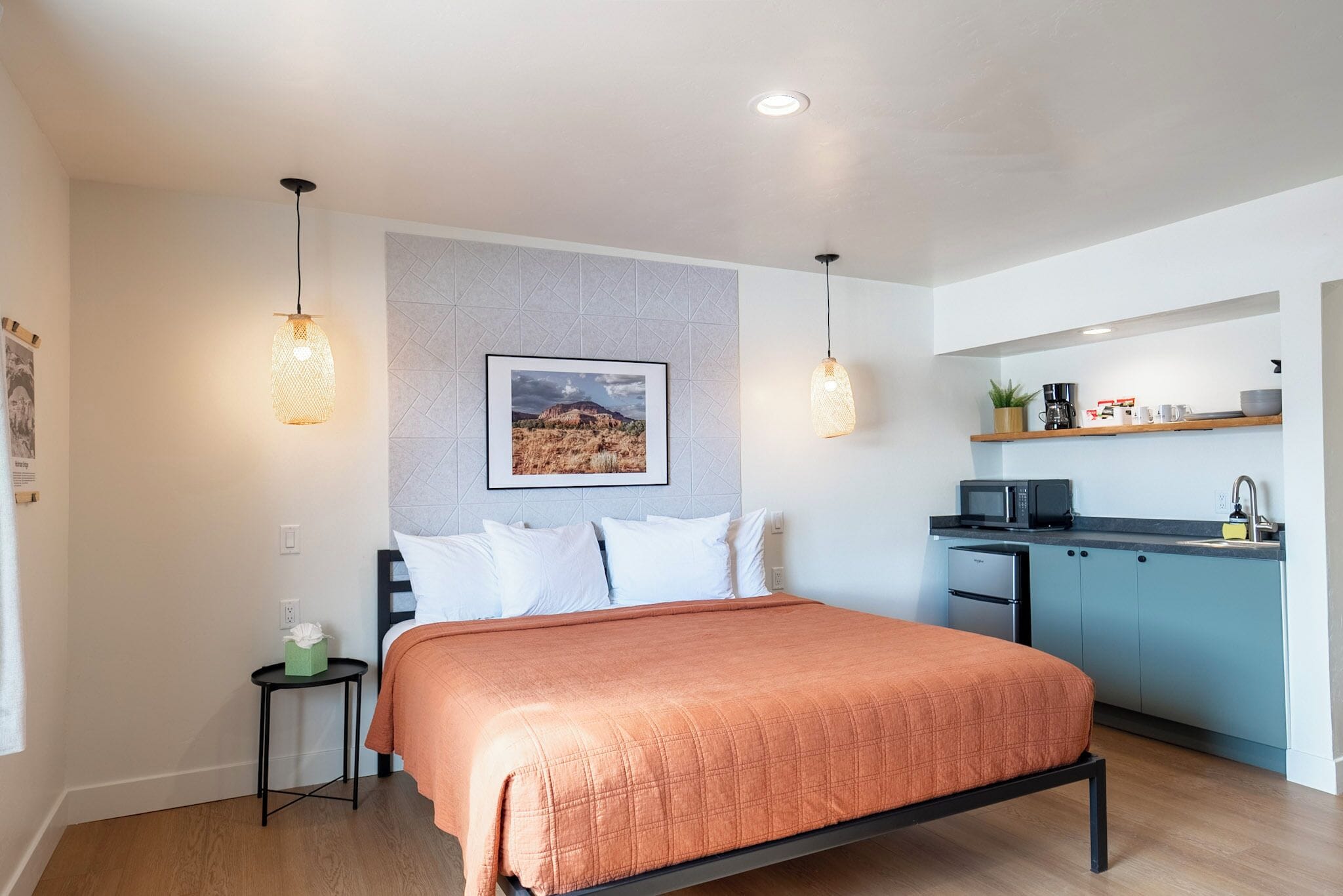 A modern bedroom with a bed, bedside table, and a small table with a plant. There is a kitchenette with a microwave and a sink.