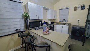 Cocina funcional con armarios blancos, microondas, tostadora y lavadora. Hay dos taburetes de bar y una planta en maceta.