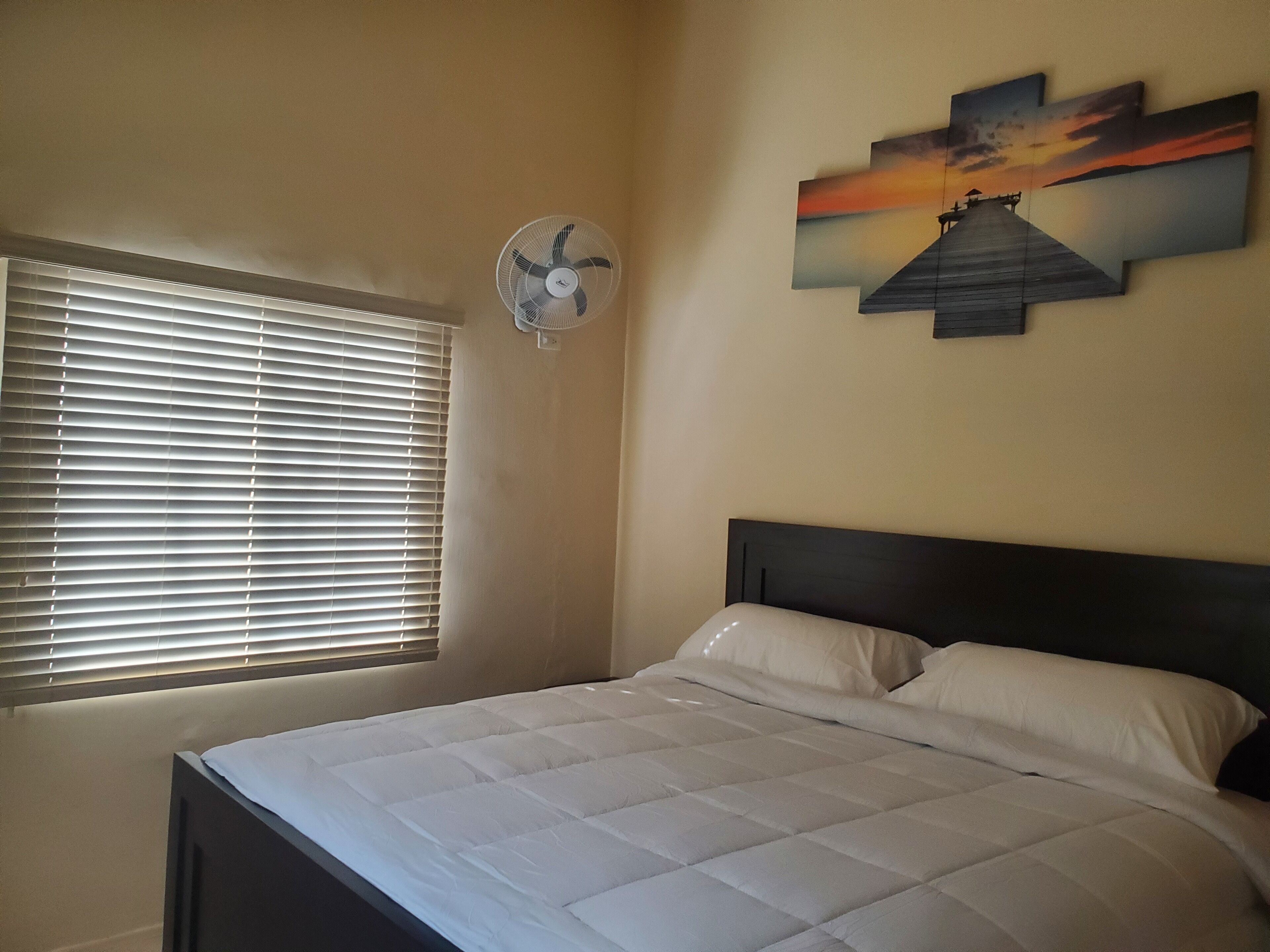 Un dormitorio con cama, ventana con persianas, ventilador de pared y una pintura de atardecer.