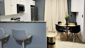 A compact kitchen with white cabinets, a microwave, and a refrigerator. There is a dining area with a round table and chairs, and a trash can.