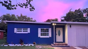 A blue house with a white door and windows, a small porch, and a brick pathway leading to the garage.