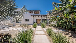 A modern house with a central pathway leading to a wooden door, surrounded by tropical plants and a clear blue sky. - Casa Alas. Newly built, private pool, and a short walk to the beach! (Todos Santos)