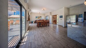 A spacious kitchen with modern appliances, a dining area with wooden chairs, and large windows offering a view of the outdoors. - Casa Alas. Newly built, private pool, and a short walk to the beach! (Todos Santos)