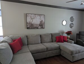 Ein graues Ecksofa mit roten Kissen, ein an der Wand befestigtes abstraktes Gemälde und dekorative sechseckige Spiegel.