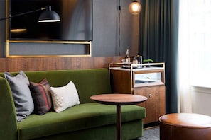 Ein grünes Sofa mit drei Kissen, ein runder Holztisch, eine kleine Holzkabine und ein an der Wand befestigter Fernseher.
